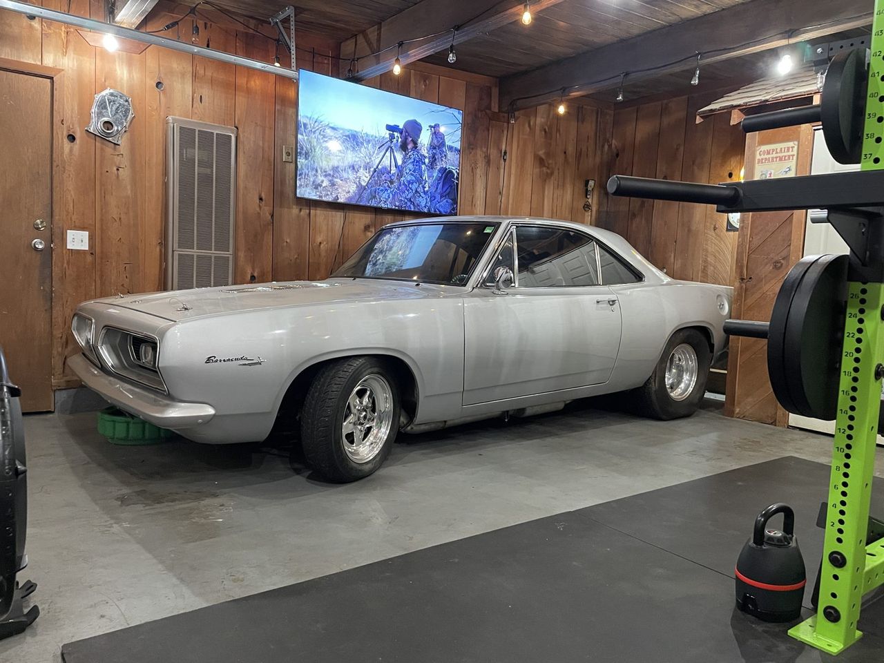 A silver 1967 Plymouth Barracuda parked in a wood-panelled home garage, sitting low on polished five-spoke wheels.