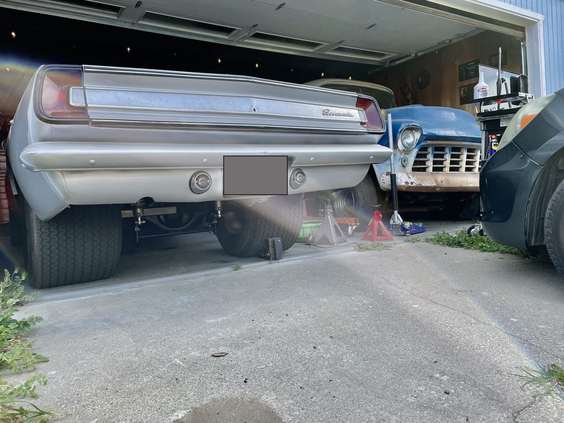 Rear view of the Barracuda in an open garage on wide rear tires, with a 1956 truck parked alongside it. The licence plate is covered.
